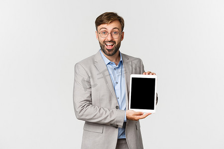 穿着灰色西装和眼镜的快乐商人的肖像，介绍应用程序或图表，显示数字平板电脑屏幕，兴奋地微笑着，站在白色背景上