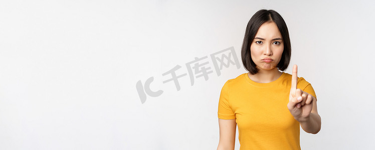 亚洲女性的肖像看起来严肃而愤怒，展示停止禁止手势、禁忌标志、禁止某物、站在白色背景上的黄色 T 恤