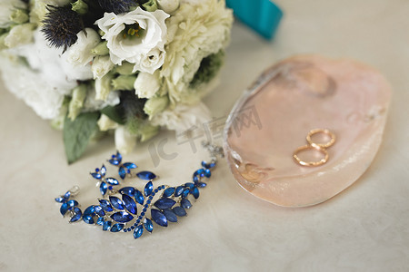 粉色大理石外壳上的金色结婚戒指，旁边是蓝色珠宝和鲜花。