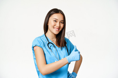 行服摄影照片_微笑的亚洲女医生、穿着磨砂服的护士展示了接种疫苗的手、流感或covid-19疫苗接种活动，竖起大拇指，白色背景