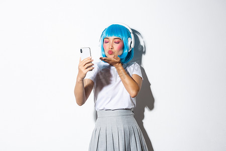 可爱的亚洲女孩打扮成动漫人物的肖像，戴着耳机和蓝色短假发自拍，用智能手机拍照，站在白色背景上