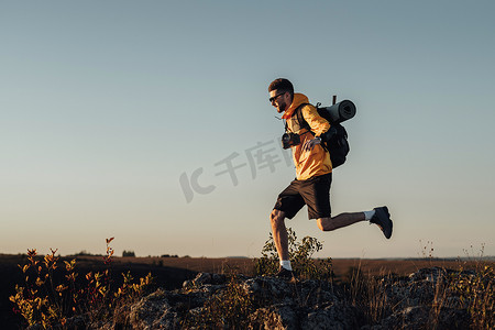 健康男性摄影照片_穿着风衣、背包和野营垫的旅行男子，在日落时跳上山顶，带数码相机的男性游牧者享受他最好的独行旅行