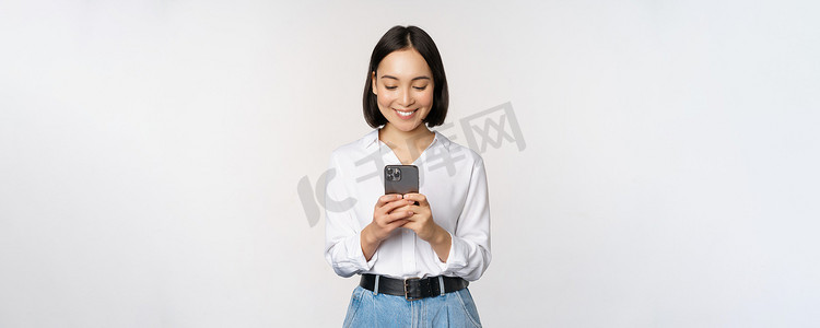 建军节手机海报摄影照片_时尚的现代亚洲女孩使用手机应用程序，用手机聊天，微笑着，站在工作室背景下的白色衬衫
