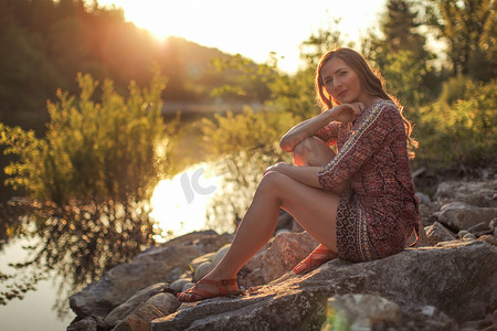 年轻女子穿着裙子，坐在河边的石头上，背景是金色的夕阳。