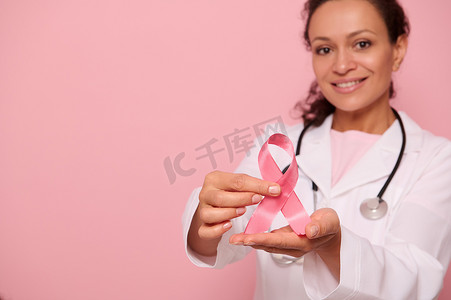关注乳腺癌意识丝带，手上是身穿白色医用外套的模糊混血女医生，隔离在粉红色背景中，复制空间。 