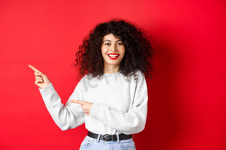 微笑的白人女性，卷发和红唇，用手指指着标志，展示促销优惠，站在工作室背景下