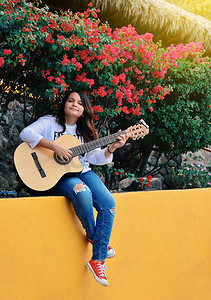 坐弹吉他摄影照片_一个女孩坐在户外弹吉他，一个微笑的女孩弹吉他的肖像，一个女孩在户外弹吉他的生活方式