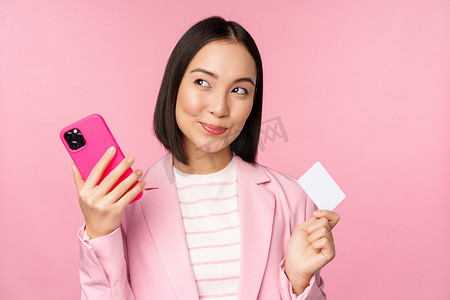 微笑的亚洲企业女性，穿着西装思考的女士，拿着智能手机和信用卡，计划在网上购买东西，用手机购物，粉红色背景