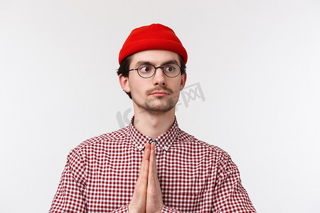 特写肖像滑稽的白人男子，留着小胡子、红色无檐小便帽和眼镜，做出合十礼或祈祷手势，表情严肃坚定，站在白色背景上