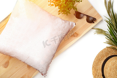 木板上有亮片的粉色枕头，旁边是黑色太阳镜、绿色植物和白色背景中突显的草帽。