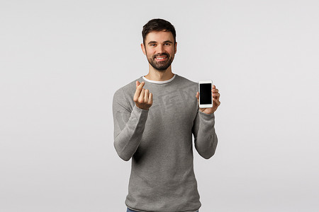 可爱而友好、迷人的留着胡子的男人使用移动应用程序玩得开心，做出韩式心形手势并拿着智能手机，展示产品、手机屏幕、站立的白色背景
