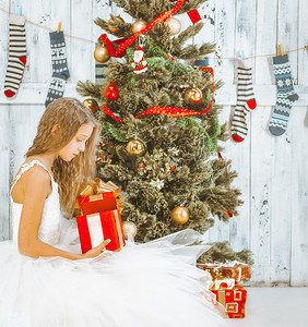身穿白色芭蕾舞短裙、长发的少女坐在圣诞树下，打开礼物。