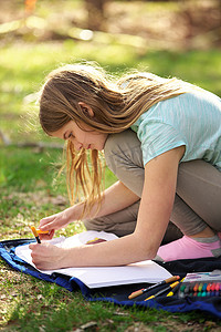 漫画美女涂色摄影照片_一个年轻的少女在草地上的毯子上的笔记本或日记中书写或涂色