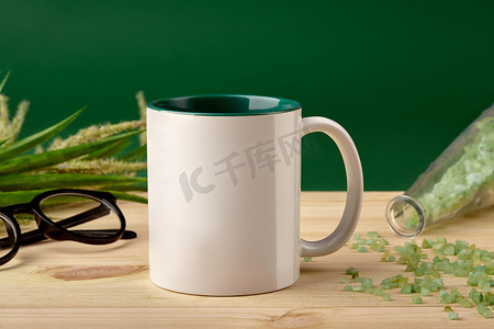 白色陶瓷杯、木质桌面上的瓶子里散落着粗海盐、玻璃杯和绿色背景上盛开的小穗。