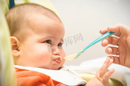 坐在高脚椅上的宝宝不肯吃妈妈用勺子给的食物，调皮、哭闹。