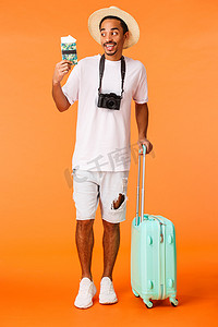 全长垂直拍摄兴奋快乐的非洲裔美国男性旅行者，迫不及待地在阳光下喝鸡尾酒，享受很棒的假期，拿着行李、手提箱和带票的护照