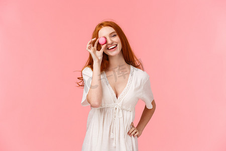 龙福袋元宝摄影照片_齐腰肖像魅力红发女性穿着白色春裙，倾斜头，无忧无虑地微笑，把马卡龙甜点放在眼睛上，享受美味的食物，站在粉红色的背景上