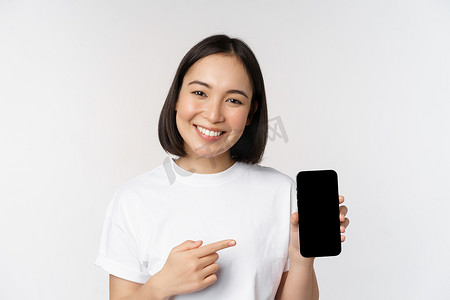 手机界面摄影照片_微笑的亚洲女性用手指指着智能手机屏幕，显示应用程序界面、手机网站，站在白色背景上