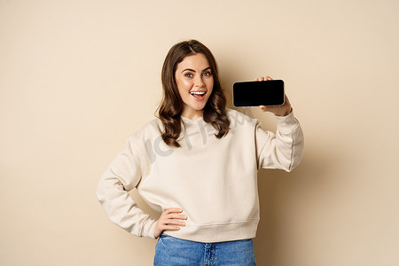时尚深色背景摄影照片_微笑的美丽女模特展示水平智能手机屏幕、移动应用程序、站在米色背景上