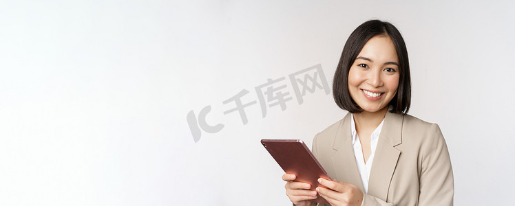 女脸手摄影照片_亚洲女商人、女售货员拿着数字平板电脑、微笑着、使用小工具、穿着西装站在白色背景上的形象
