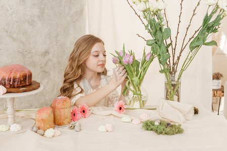 一个穿着浅色连衣裙、长发的女孩坐在复活节餐桌旁，桌上摆着蛋糕、春天的花朵和鹌鹑蛋。