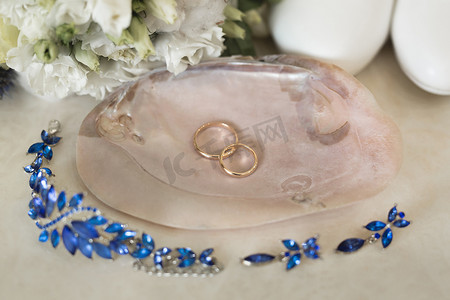 粉色大理石外壳上的金色结婚戒指，旁边是蓝色珠宝和鲜花。