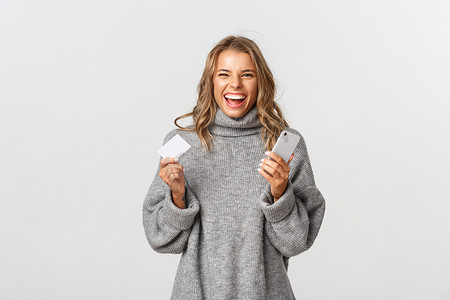 快乐的金发女孩拿着智能手机和信用卡，享受网上购物，站在白色背景下的形象