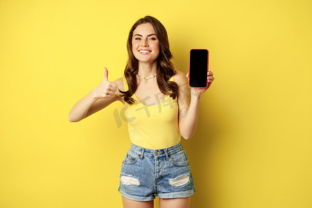 快乐微笑的时尚女孩竖起大拇指和智能手机应用程序界面、手机显示屏，对网站、在线购物应用程序感到满意，站在黄色背景上