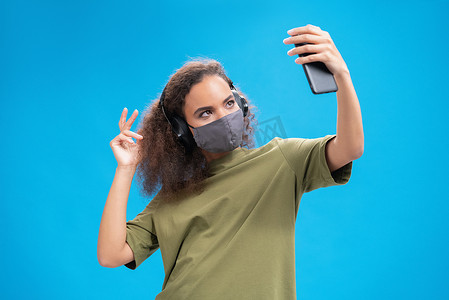 跳舞的非洲裔美国女孩在智能手机上听音乐，戴着橄榄色T恤的耳机和可重复使用的面罩，以防止其他人感染蓝色背景下隔离的电晕COVID-19感染