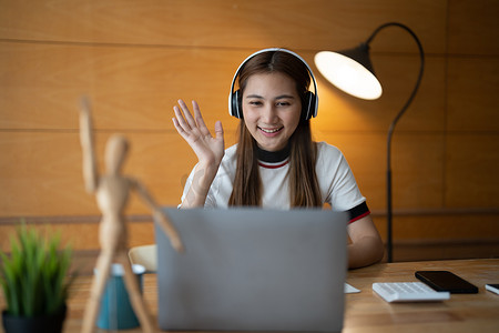 戴耳机的亚洲年轻女性微笑着迎接在笔记本电脑上的网络摄像头虚拟对话中交谈，戴无线耳机的快乐女性在电脑上进行视频通话，在线咨询客户