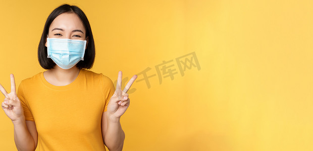 冠状病毒表现摄影照片_积极的亚洲女性微笑着，在大流行期间戴着covid-19医用口罩，表现​​出和平V手势，黄色背景