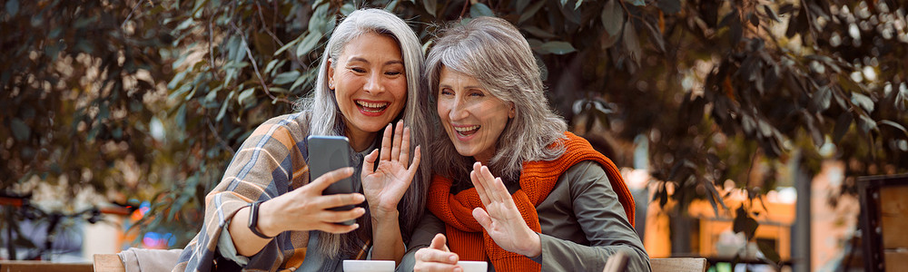 积极的成熟女性朋友在街头咖啡馆通过智能手机进行视频聊天时挥手