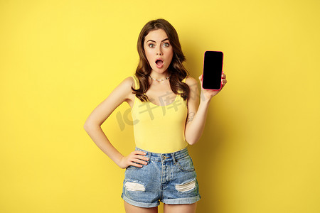 背心摄影照片_时尚的微笑女性展示手机屏幕、智能手机上的应用程序界面、站在背心和黄色背景的短裤
