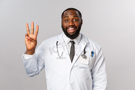 三个点摄影照片_英俊快乐的非洲裔美国男医生的腰部肖像，穿白大褂的医生，展示三个手指，点东西或要求护士带第三个病人进来，灰色背景