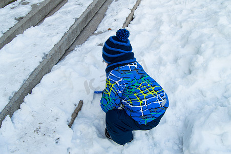 冬天小男孩摄影照片_冬天，一个戴着蓝色针织帽和一件温暖夹克的小男孩在覆盖着雪的楼梯附近玩耍，从后面看