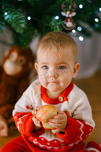 小孩眼睛摄影照片_穿着针织圣诞服装的可爱小孩在圣诞树前拿着一个苹果