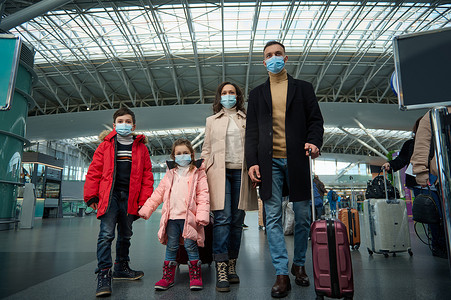 一个年轻家庭戴着防护性医用口罩在大流行期间旅行的肖像，在机场等待办理登机手续时提着行李和手提箱站在机场。