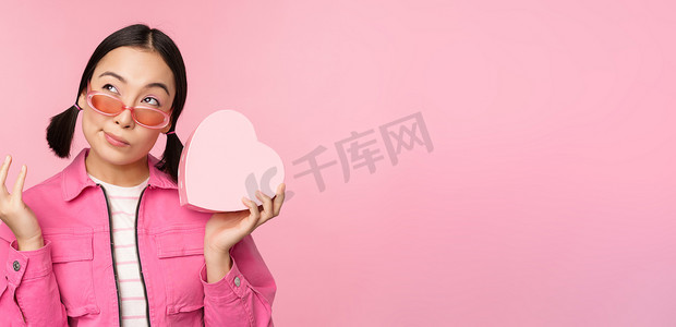 戴着太阳镜的时尚亚洲女友的形象，猜猜礼盒里有什么，心形礼物，站在粉色背景上