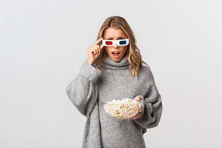 戴着 3D 眼镜的金发女孩，拿着爆米花碗，看电影，站在白色背景上，看上去很困惑