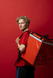 挂着的包摄影照片_身穿红色制服 T 恤、肩上挂着红色食品保温袋的年轻快递员站在红色背景工作室里。
