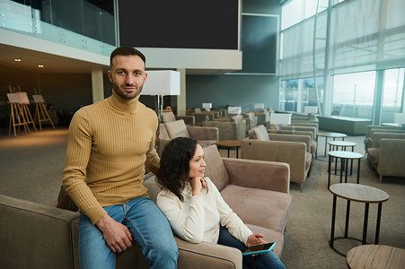 英俊迷人的白人男子看着镜头拥抱他的女朋友，坐在国际机场休息室的椅子上，等待登机。