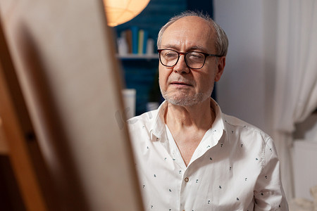 男老人头发摄影照片_退休男艺术家在用铅笔绘画时在画架上的画布上寻找灵感的特写肖像
