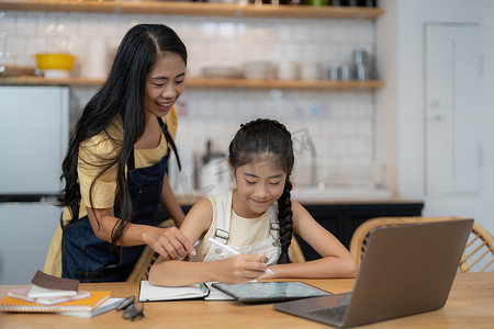 学生写做作业摄影照片_亚洲母亲和小女儿坐在厨房的桌子上一起在线学习，混血妈妈和小女孩手写，做作业，在家学习，在家上学的概念