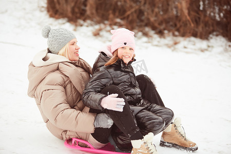 快乐的母亲带着女婴坐在雪橇上，从山上的雪上滑下来。