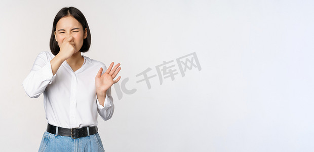 女表摄影照片_厌恶的亚洲女孩闭着鼻子，表现出拒绝的姿态，恶心难闻的气味，站在白色背景上