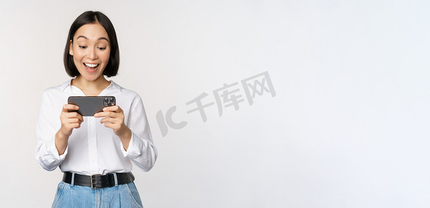 建军节手机海报摄影照片_年轻的亚洲女性在智能手机应用程序上观看、手持手机、看着屏幕、笑着、站在白色背景上的形象