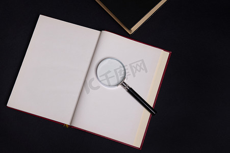 放大镜的平躺组合物在一本打开的书上，硬红色封面，隔离在黑色背景上，有文本空间。