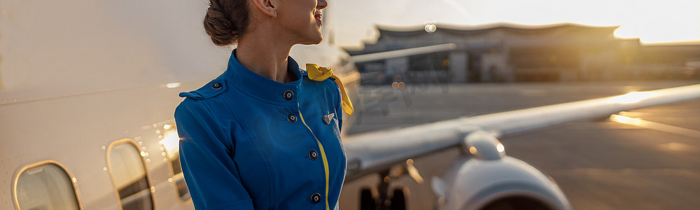 穿着蓝色制服的体贴的空姐看着别处，站在户外夕阳下。