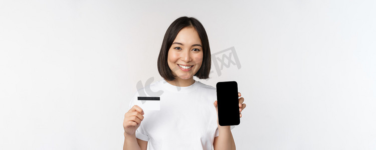站在白色背景上微笑的年轻亚洲女性的肖像，展示手机屏幕和信用卡