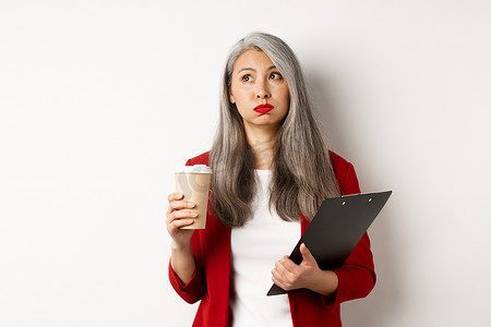 女性疲惫摄影照片_疲惫的亚洲女上班族拿着剪贴板和纸杯，喝着咖啡，面容疲惫地呼气，站在白色背景上